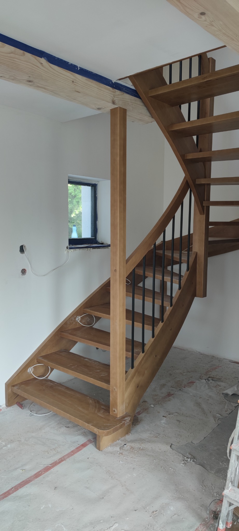 Wewnętrzne kręte schody z jasnego drewna z czarnymi metalowymi prętami balustrady, widok na okno i surowy stan wykończenia wnętrza.