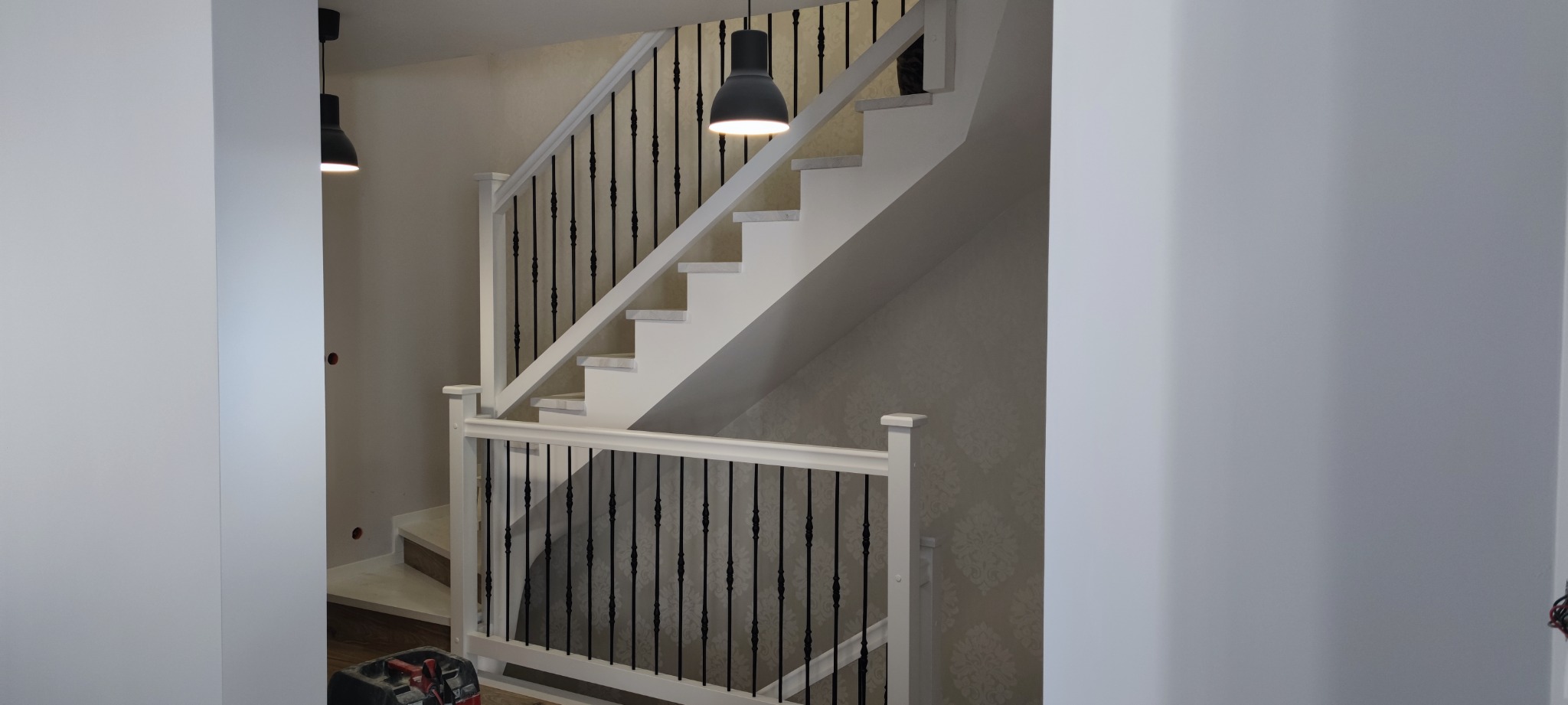 Białe schody z czarnymi balustradami w nowoczesnym domu, widoczna skrzynka narzędziowa na dole.