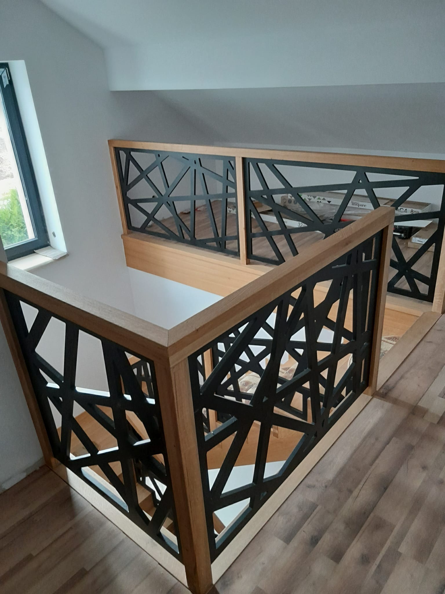 Nowoczesna balustrada z jasnego drewna i czarnych, geometrycznych wypełnień na schodach w jasnym wnętrzu z oknem dachowym.