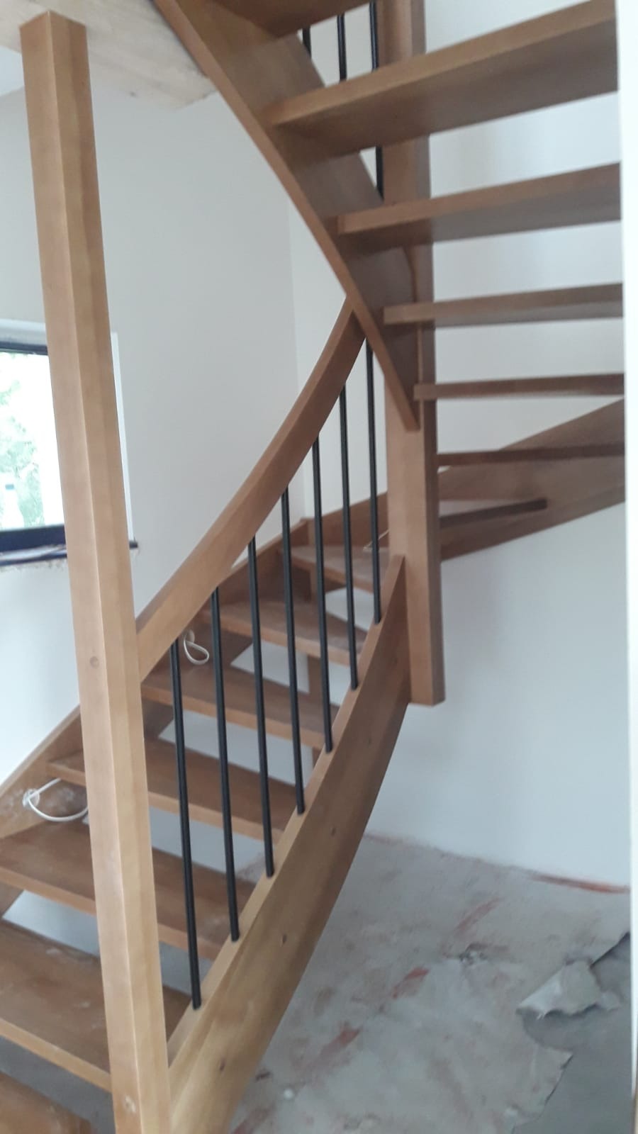 Wewnętrzne schody z jasnego drewna z czarnymi, metalowymi balustradami, widok od dołu, w tle okno i surowa podłoga w trakcie remontu.