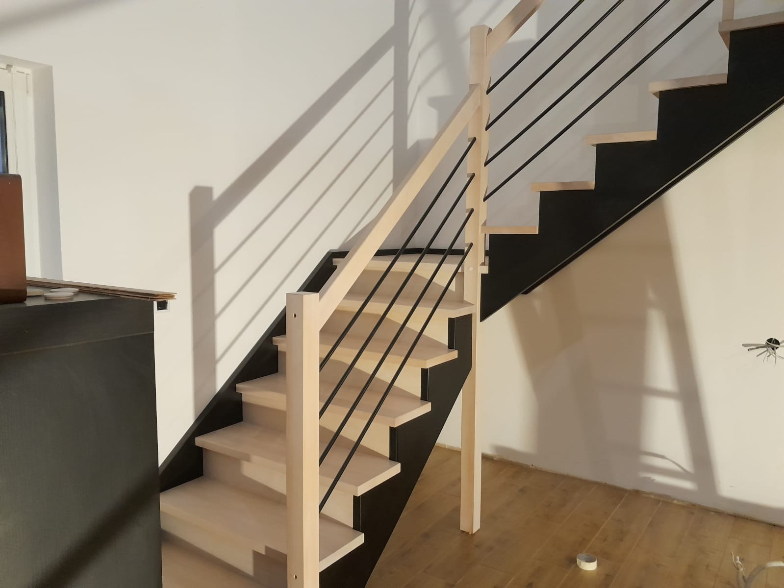 Nowoczesne schody z jasnego drewna z czarną konstrukcją i metalowymi, czarnymi balustradami, widoczne na tle drewnianej podłogi i białej ściany w trakcie montażu.
