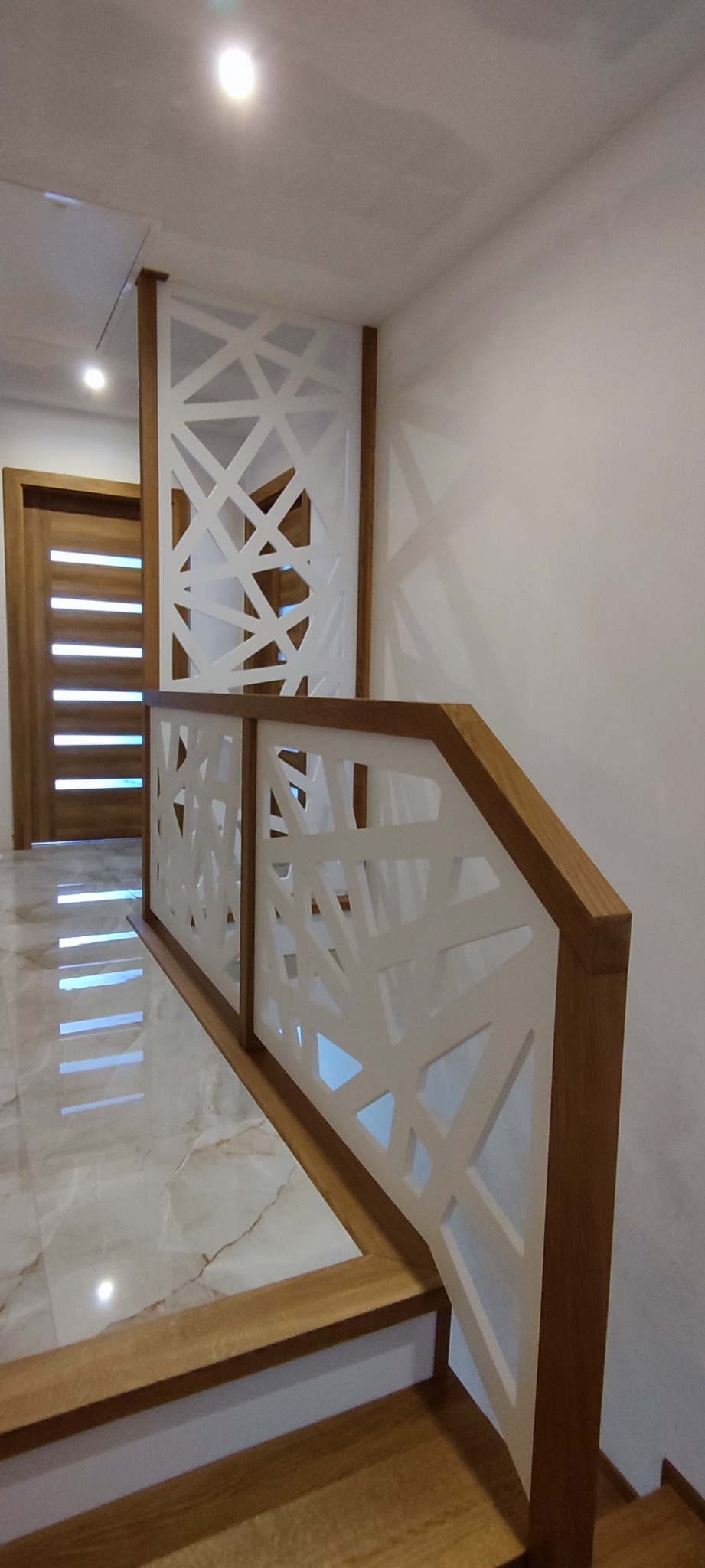 Nowoczesna balustrada schodowa z drewnianą ramą i białym, ażurowym wypełnieniem o geometrycznym wzorze, widoczna również część schodów i drzwi wejściowe w tle.