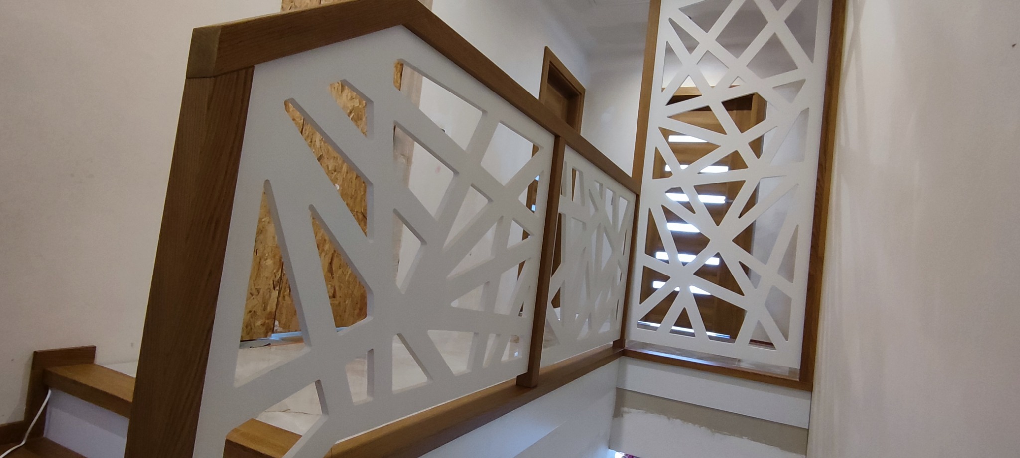 Nowoczesna balustrada schodowa z białymi, ażurowymi panelami o geometrycznym wzorze i drewnianymi ramami, widok z dołu na schody.