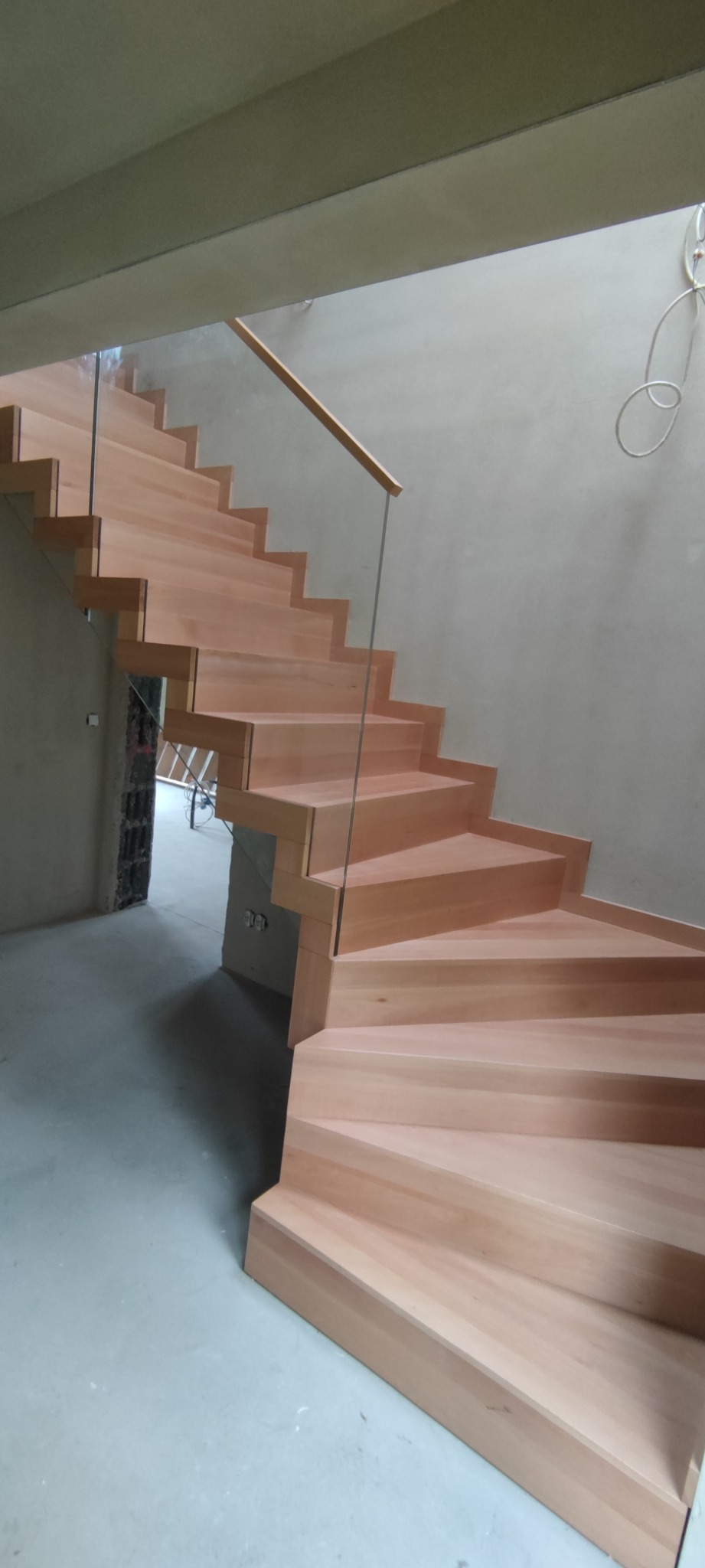 Wewnętrzne schody z jasnego drewna z przezroczystą szklaną balustradą zamontowane w surowym stanie budynku, widoczne niedokończone ściany i podłogi.