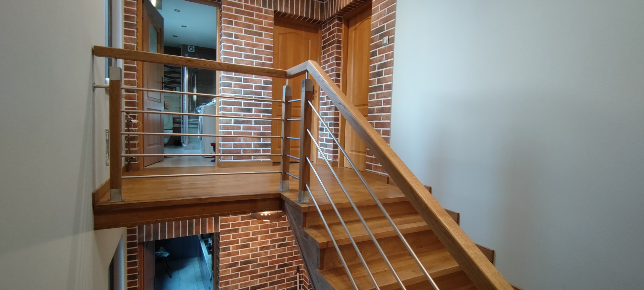 Drewniane schody z metalową balustradą na tle ściany z cegły klinkierowej i drewnianych drzwi.