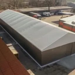 Widok z g&oacute;ry na długą, jednopiętrową halę magazynową z szarą, trapezową blachą i białym dachem, stojącą obok innych budynk&oacute;w przemysłowych i placu z zaparkowanymi ciężar&oacute;wkami.