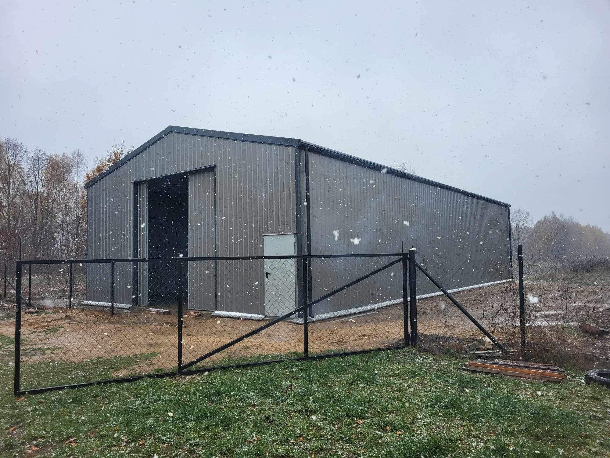 Szara hala z otwartymi przesuwnymi drzwiami i białymi drzwiami wejściowymi, otoczona czarnym ogrodzeniem z siatki w zimowej scenerii z padającym śniegiem.