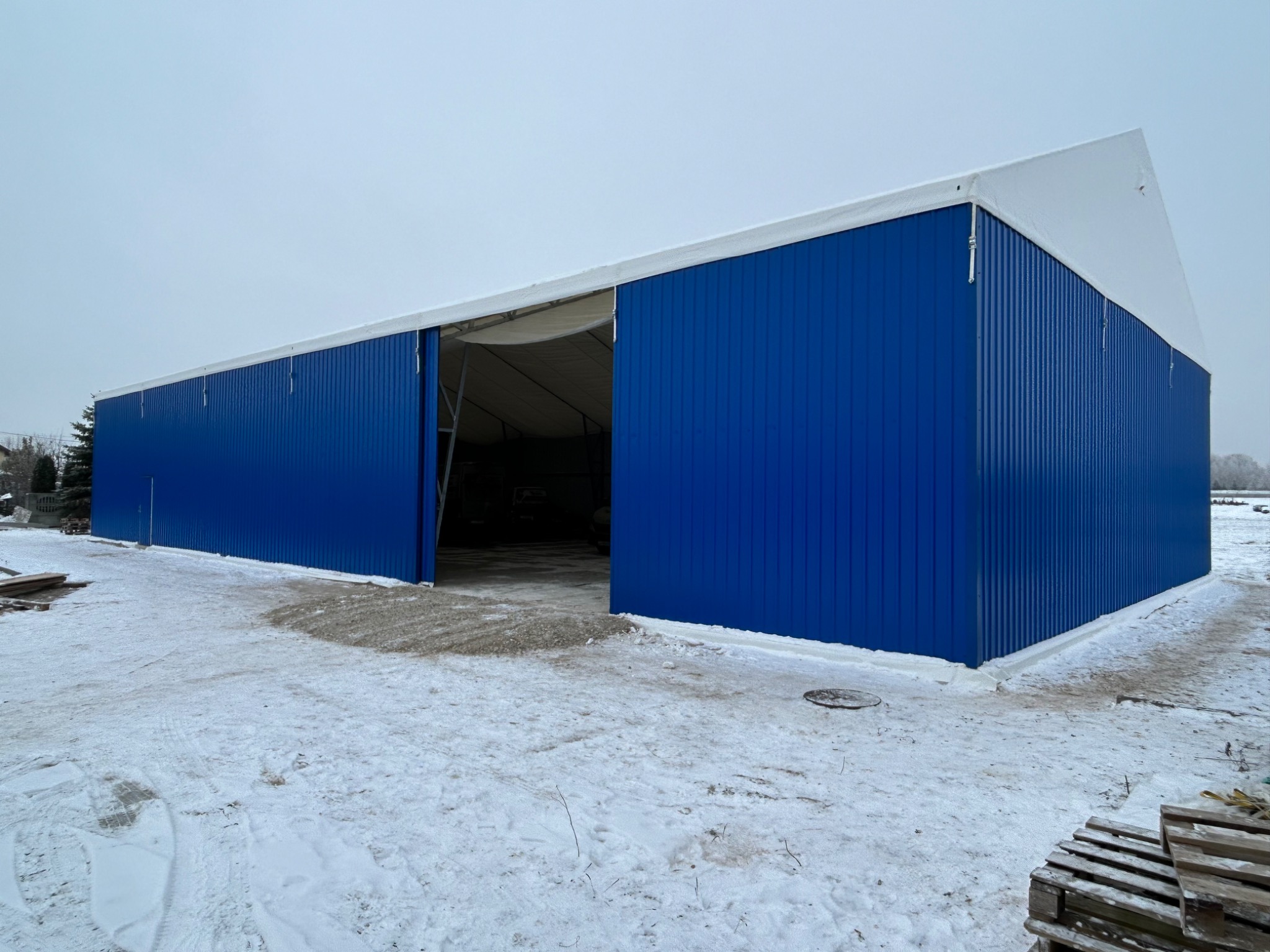 Długa, niebieska hala z blachy falistej z białym dachem w zimowym krajobrazie, częściowo otwarte przesuwne wrota.
