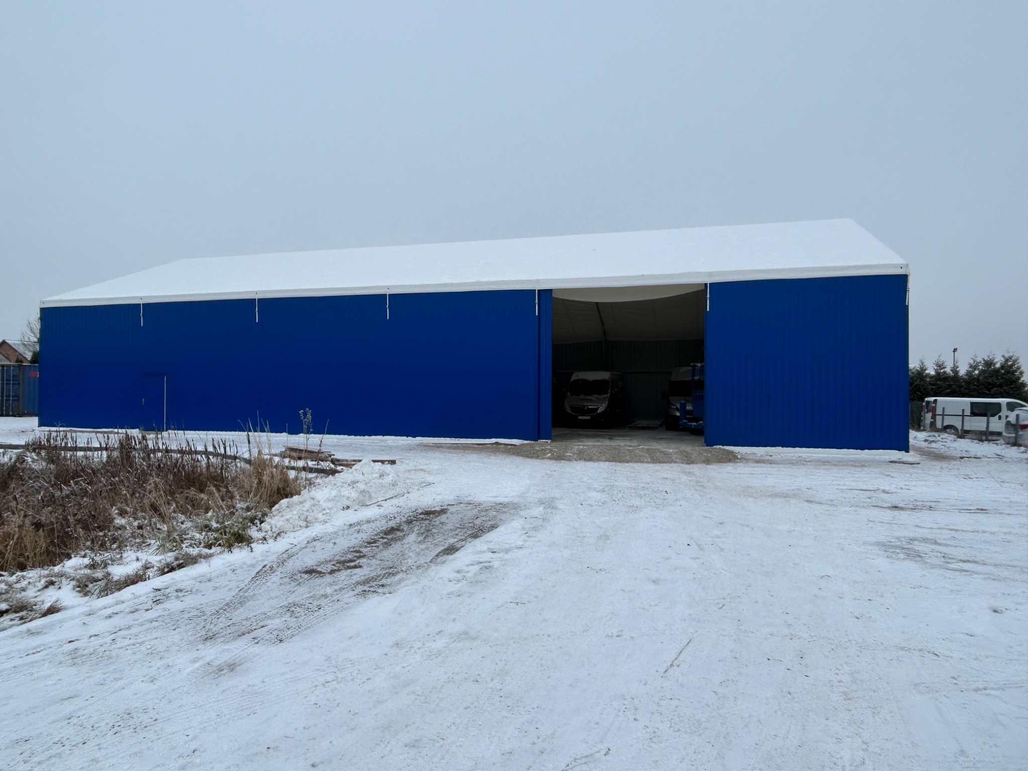 Zimowy widok na dużą, niebieską halę z białym dachem, z otwartymi wrotami, w których widać zaparkowane pojazdy dostawcze.