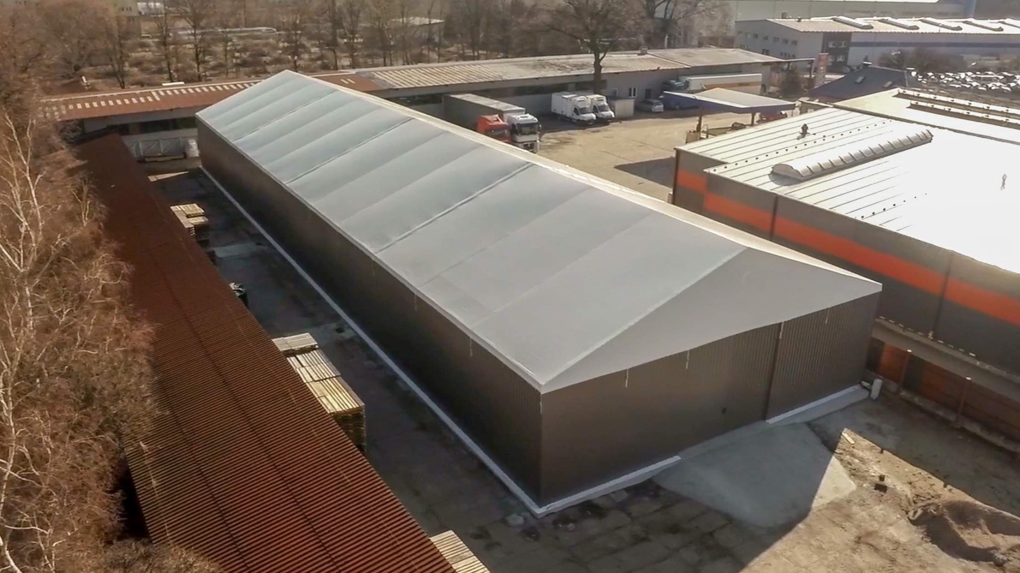 Widok z góry na długą, jednopiętrową halę magazynową z szarą, trapezową blachą i białym dachem, stojącą obok innych budynków przemysłowych i placu z zaparkowanymi ciężarówkami.
