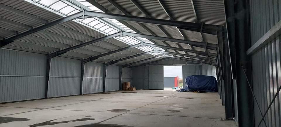 Wnętrze hali z metalową konstrukcją szkieletową, blachą trapezową na ścianach i dachu z świetlikami, betonowa posadzka, częściowo otwarte wrota w tle.
