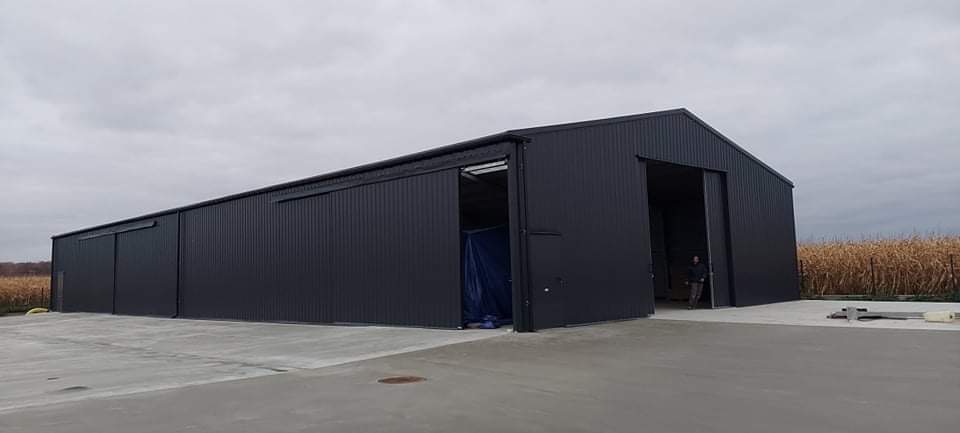 Czarna hala magazynowa z przesuwnymi drzwiami, widoczna osoba wewnątrz, betonowy plac przed halą, w tle pole z zaschniętą roślinnością.