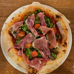 Pizza z szynką parmeńską, rukolą i pomidorkami koktajlowymi na talerzu, z widocznym brzegiem ciasta z charakterystycznymi przypaleniami.