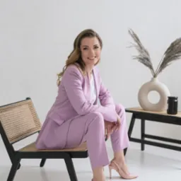 Uśmiechnięta kobieta w liliowym garniturze siedzi na designerskim krześle z rattanu na tle minimalistycznego wnętrza z beżową ceramiką i suszonymi trawami.