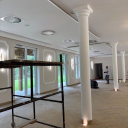 Adam Bujnowski - Przestronne wnętrze sali bankietowej z kolumnami i drewnianą podłogą w jodełkę. Widoczne rusztowanie malarskie i pracownik sprzątający podłogę.