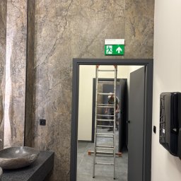 Adam Bujnowski - Łazienka w trakcie wykończenia: marmurowe ściany, kamienna umywalka, czarne drzwi i drabina. Widoczny pracownik w głębi. Oświetlenie sufitowe.