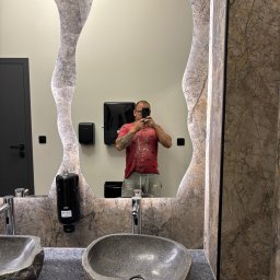 Adam Bujnowski - Wykończona łazienka z kamienną umywalką i lustrem o nieregularnym kształcie, odbicie malarza w ubraniu roboczym. Detale marmurowej okładziny i nowoczesna armatura.