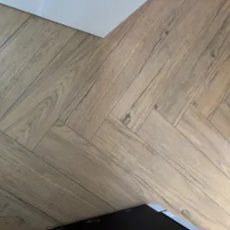 Parkiet ułożony w jodełkę francuską, z widoczną strukturą drewna i subtelnymi odcieniami brązu. Ujęcie z góry, z fragmentem białej ściany i czarnego przedmiotu w rogu.