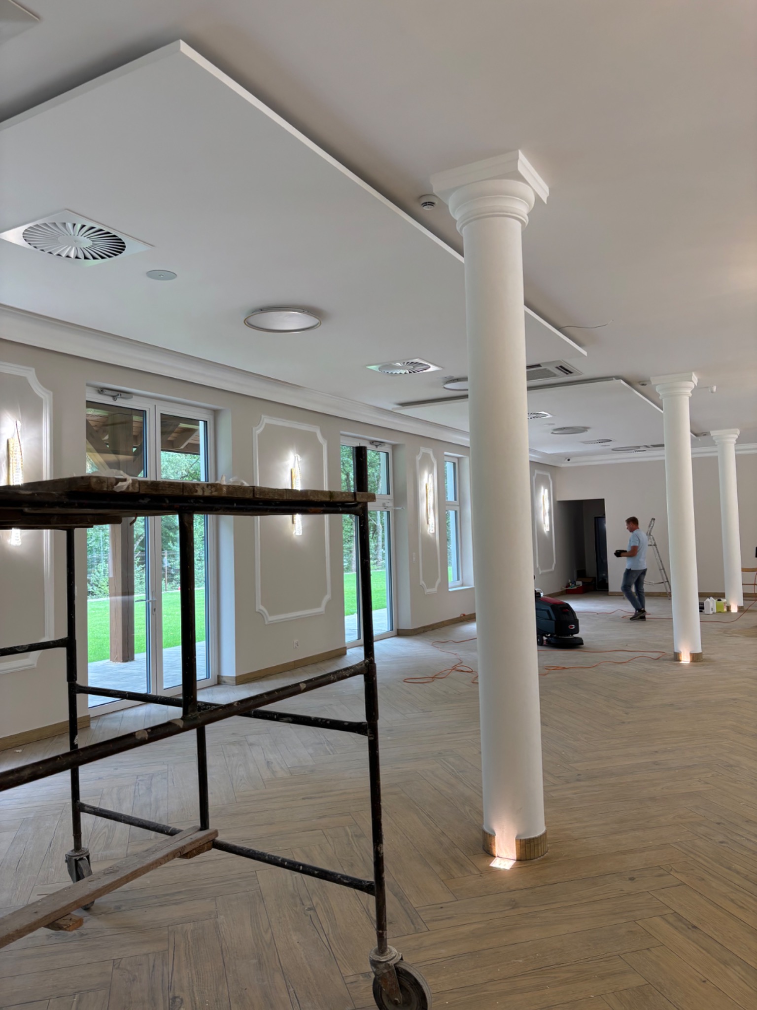 Przestronne wnętrze sali bankietowej z kolumnami i drewnianą podłogą w jodełkę. Widoczne rusztowanie malarskie i pracownik sprzątający podłogę.