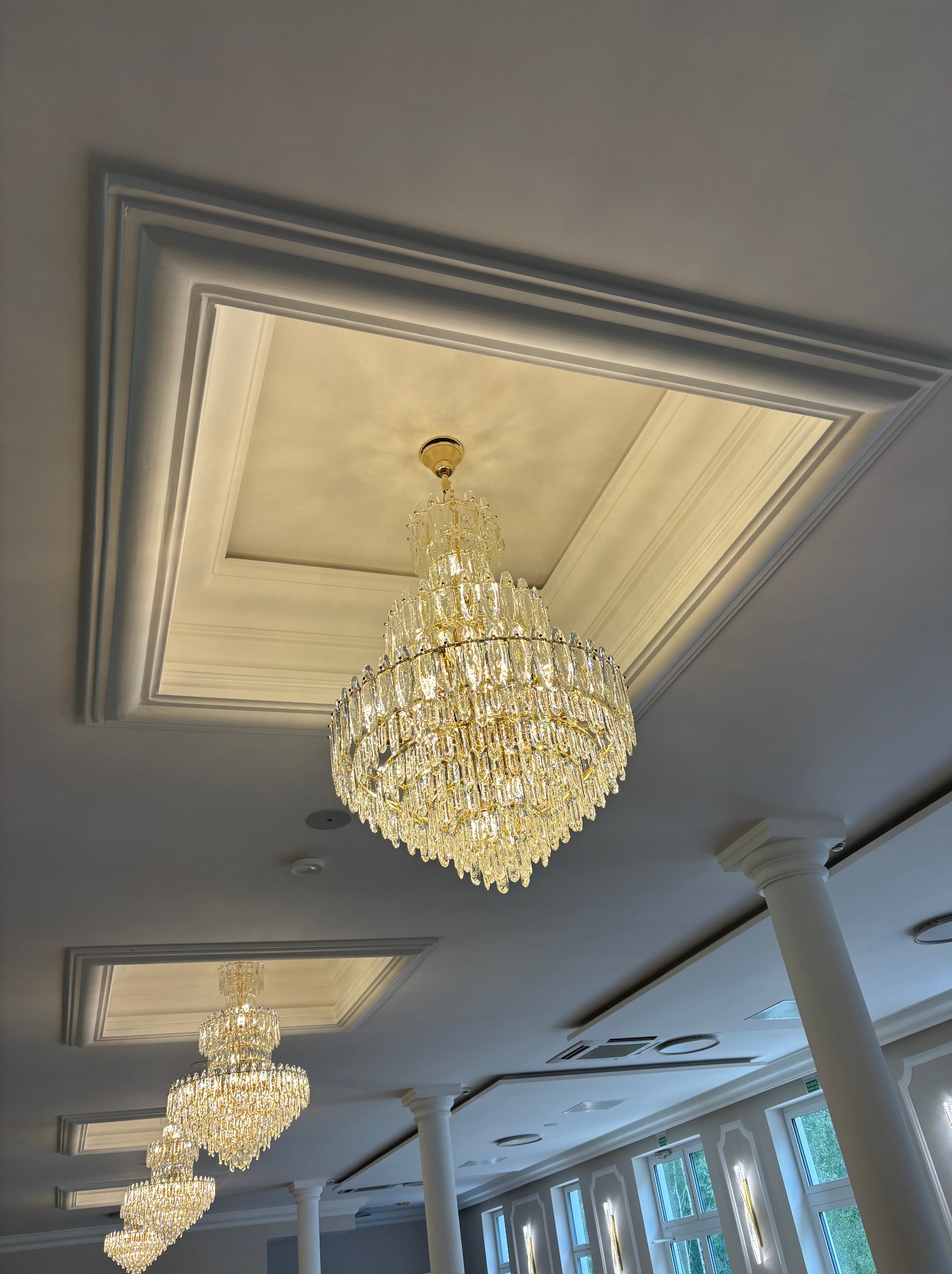 Kryształowy żyrandol w suficie z dekoracyjnymi listwami. Widok z dołu na eleganckie oświetlenie i detale wykończeniowe sufitu w jasnym wnętrzu.