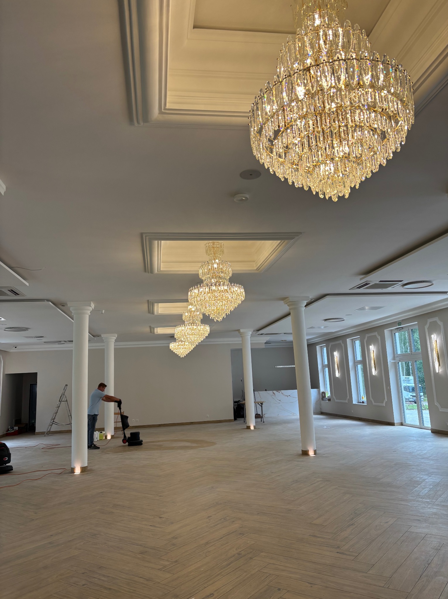 Polerowanie parkietu ułożonego w jodełkę w przestronnym, jasnym wnętrzu z kolumnami i kryształowymi żyrandolami. Pracownik używa maszyny do polerowania.