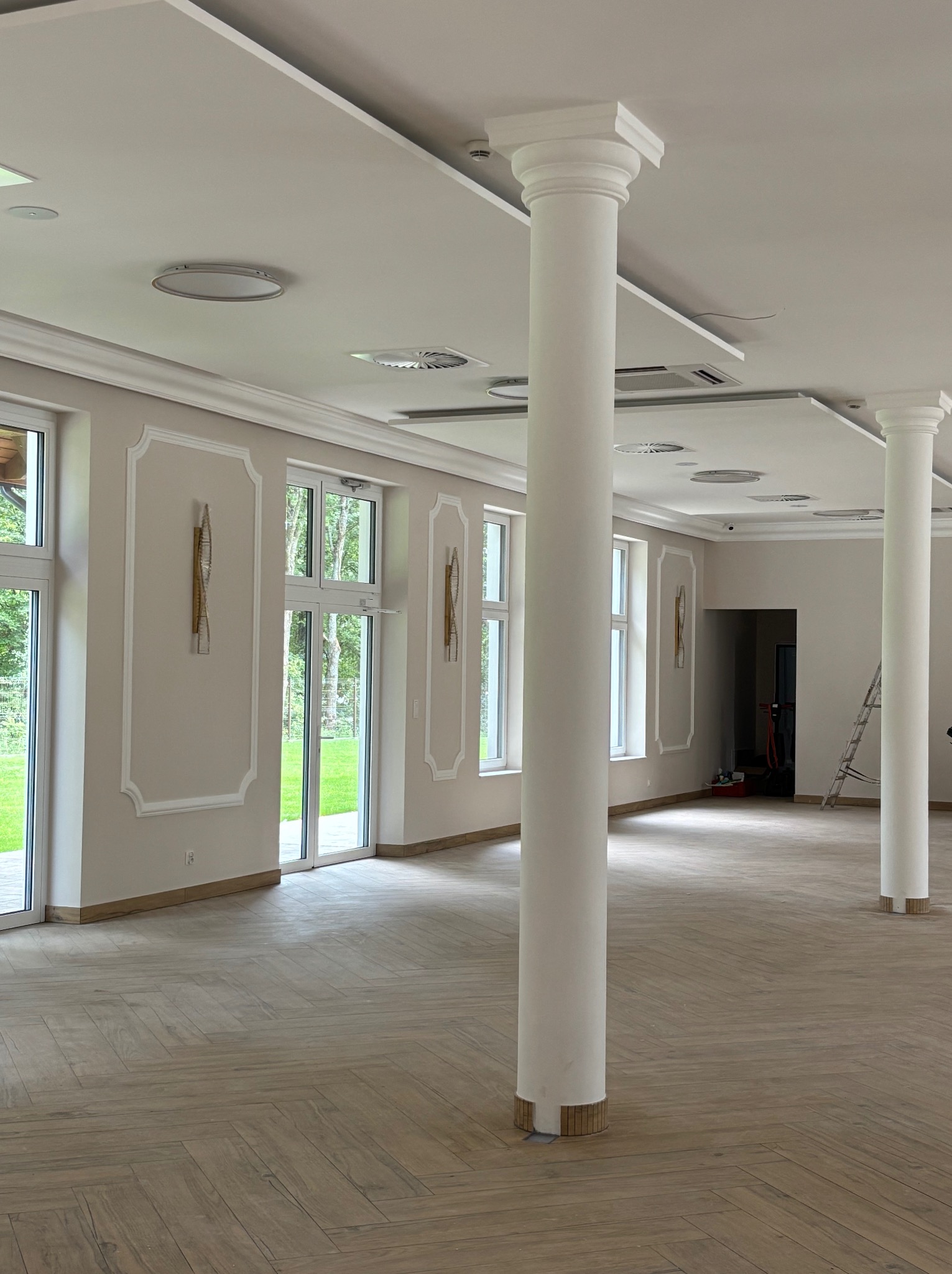 Jasne, przestronne wnętrze po remoncie z kolumnami, parkietem ułożonym w jodełkę i sztukaterią. Widoczne okna z widokiem na zieleń i drabina w tle.