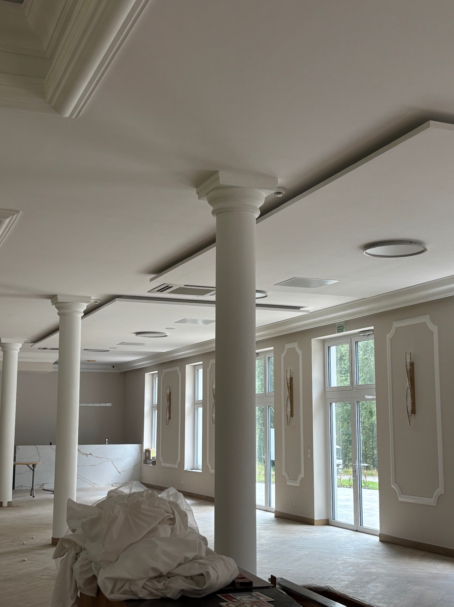 Jasne, przestronne wnętrze z kolumnami i sztukaterią, po remoncie. Widoczne wykończenia sufitu i ścian, duże okna z dekoracyjnymi obramowaniami. Podłoga w trakcie prac.