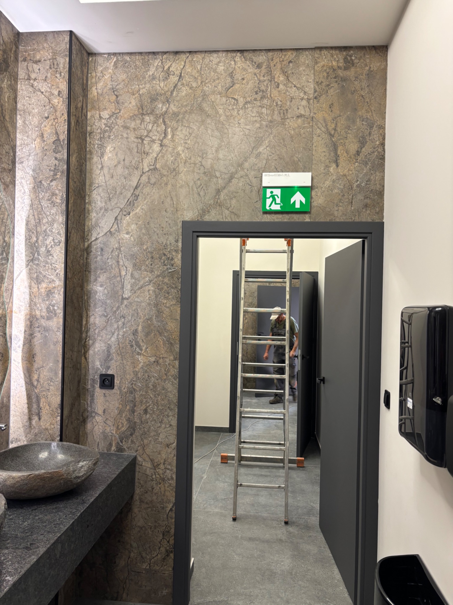 Łazienka w trakcie wykończenia: marmurowe ściany, kamienna umywalka, czarne drzwi i drabina. Widoczny pracownik w głębi. Oświetlenie sufitowe.