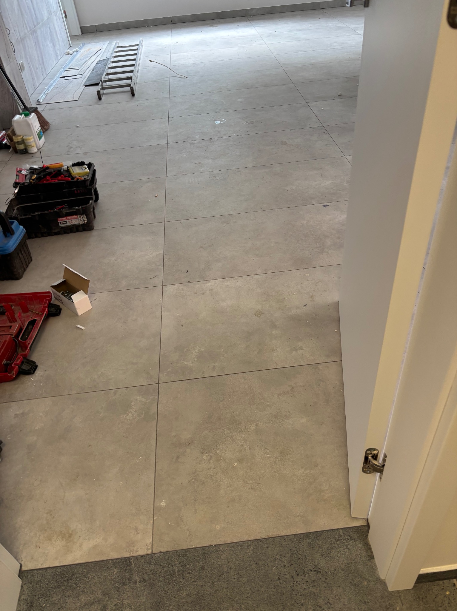 Wnętrze w trakcie remontu: świeżo ułożona, szara podłoga z płytek, narzędzia pracy i drabina w tle. Widoczny fragment białych drzwi.