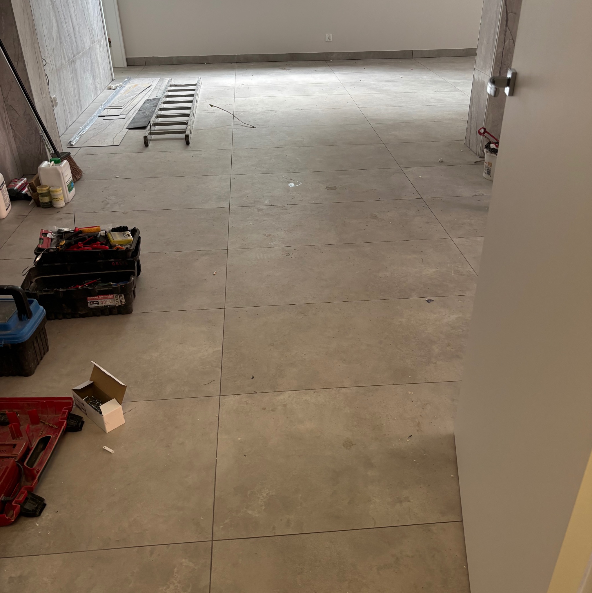 Wnętrze w trakcie remontu z narzędziami, drabiną i materiałami budowlanymi. Widoczne płytki podłogowe i częściowo wykończone ściany.