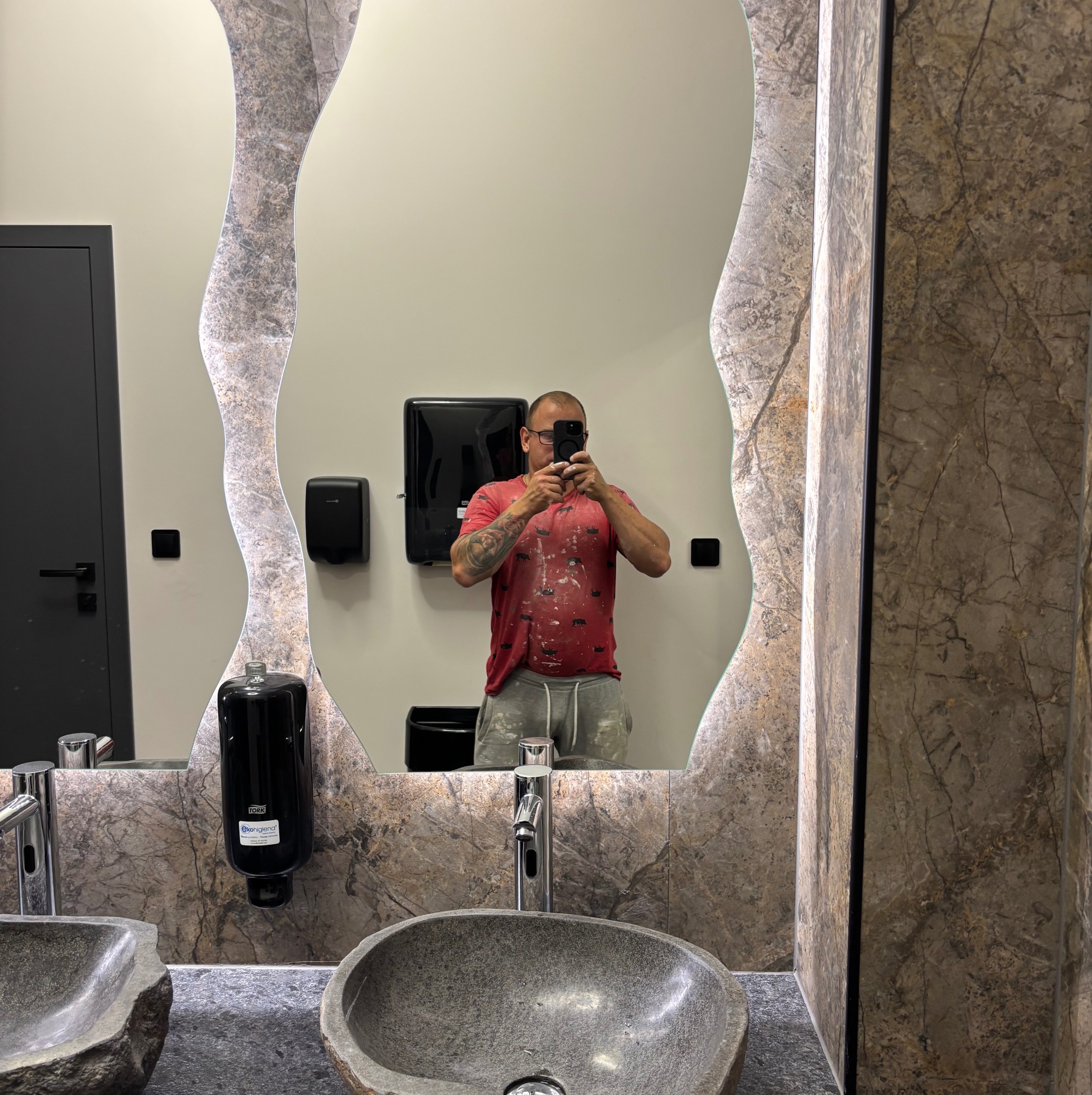 Wykończona łazienka z kamienną umywalką i lustrem o nieregularnym kształcie, odbicie malarza w ubraniu roboczym. Detale marmurowej okładziny i nowoczesna armatura.