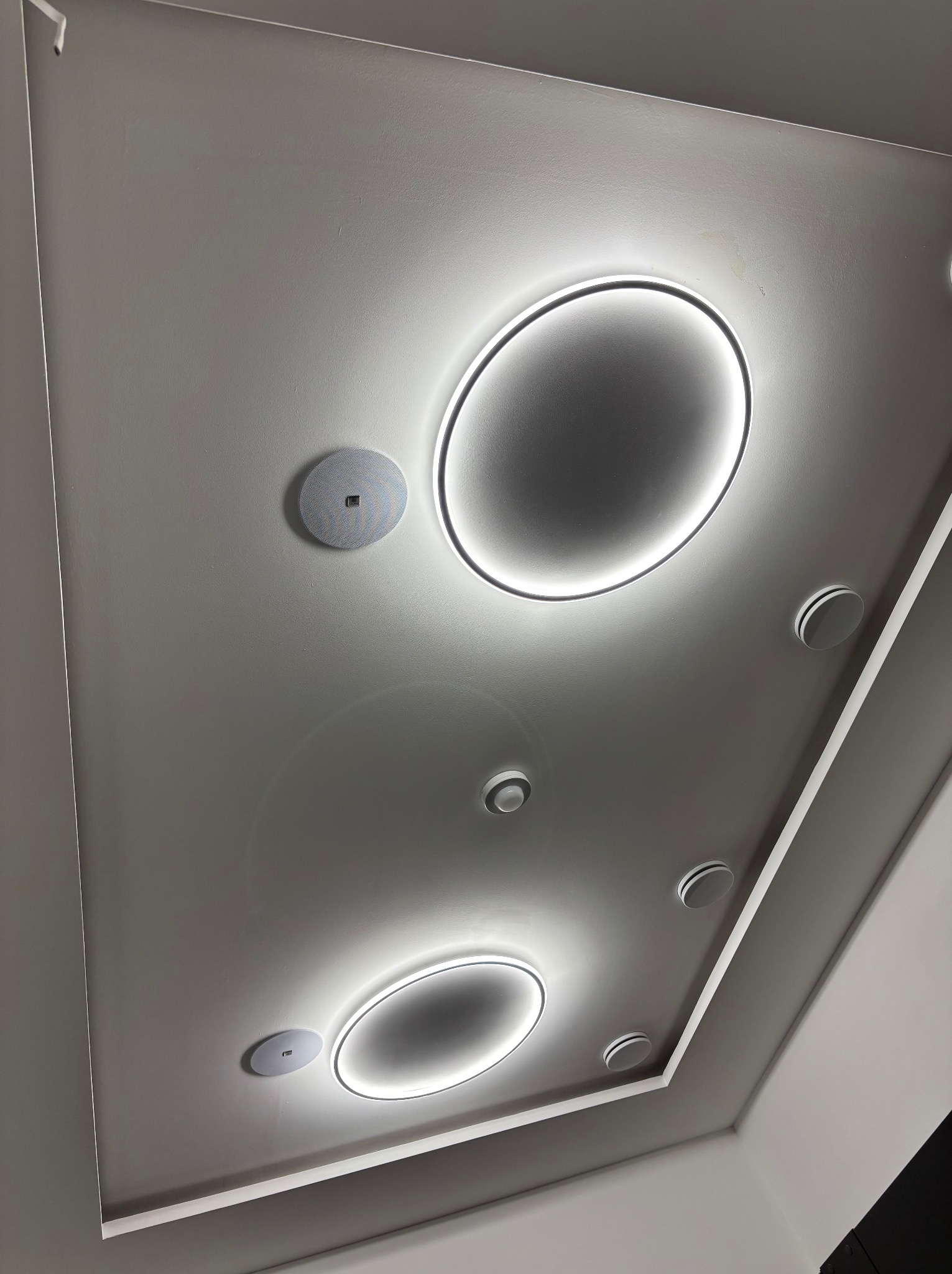 Nowoczesny sufit z podwieszanym oświetleniem LED w kształcie okręgów i punktowymi lampami. Minimalistyczny design, białe wykończenie, skosy sufitowe.