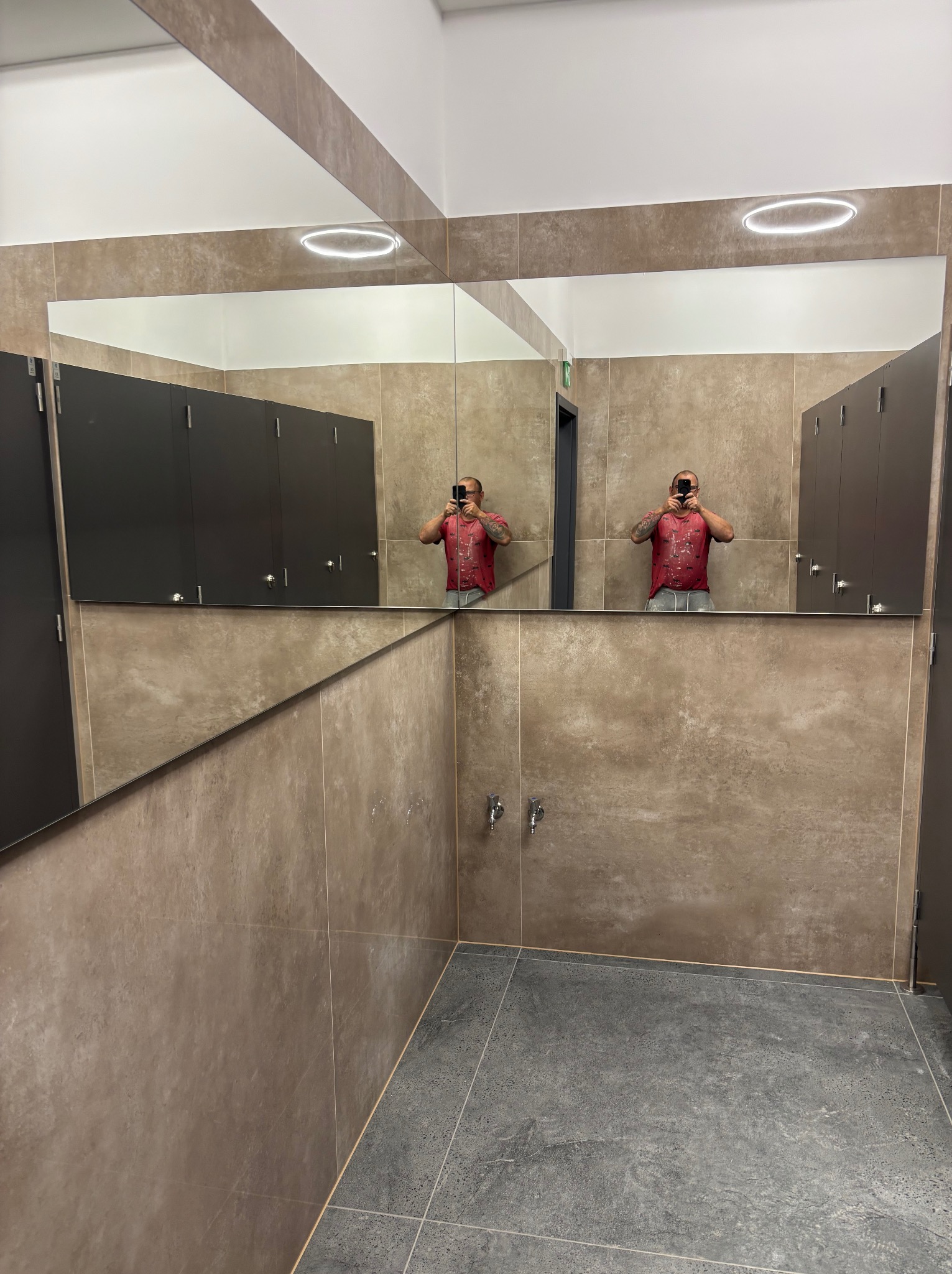 Nowoczesna łazienka publiczna z dużym lustrem, szarymi płytkami podłogowymi i brązowymi płytkami ściennymi. W lustrze odbija się osoba robiąca zdjęcie.
