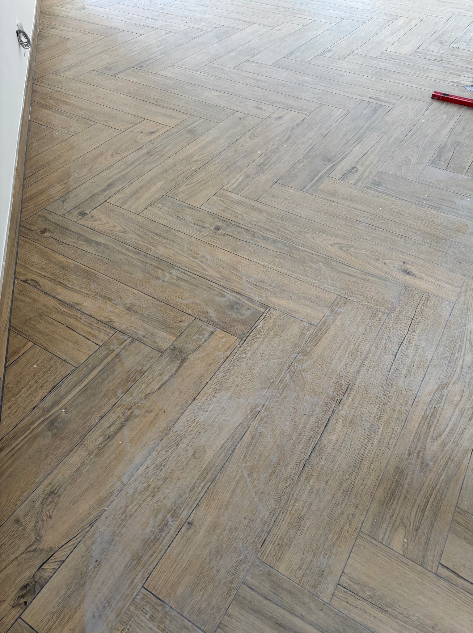 Ułożona podłoga z płytek imitujących drewno w jodełkę, widoczne fugi i fragment poziomicy. Jasne wnętrze, ukończony etap prac wykończeniowych.