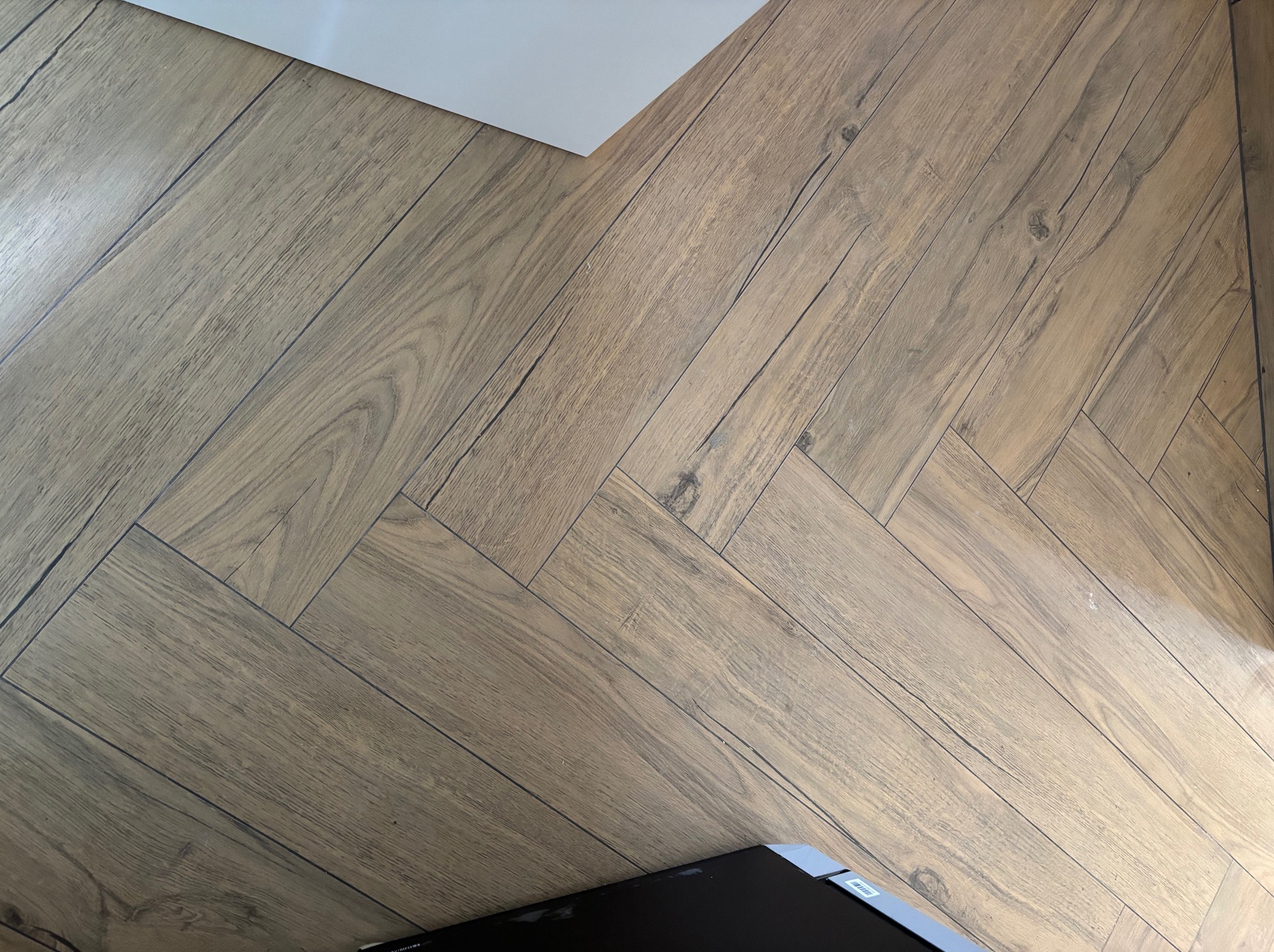 Parkiet ułożony w jodełkę francuską, z widoczną strukturą drewna i subtelnymi odcieniami brązu. Ujęcie z góry, z fragmentem białej ściany i czarnego przedmiotu w rogu.