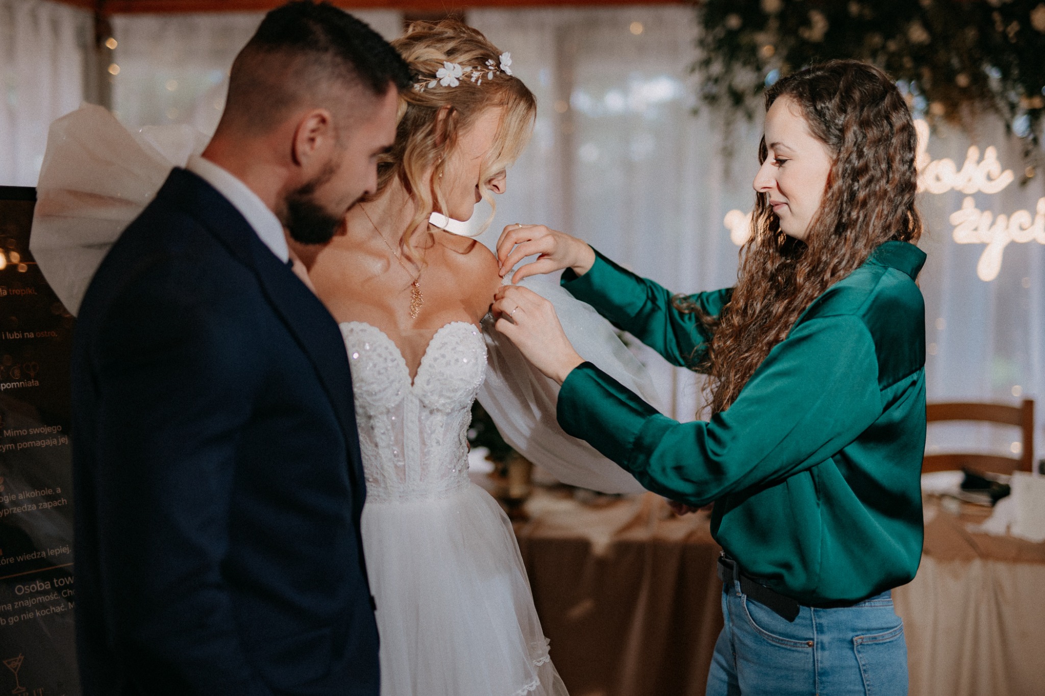 Kobieta w szmaragdowej bluzce poprawia suknię ślubną panny młodej, obok stoi mężczyzna w garniturze, w tle dekoracje ślubne i napis świetlny.