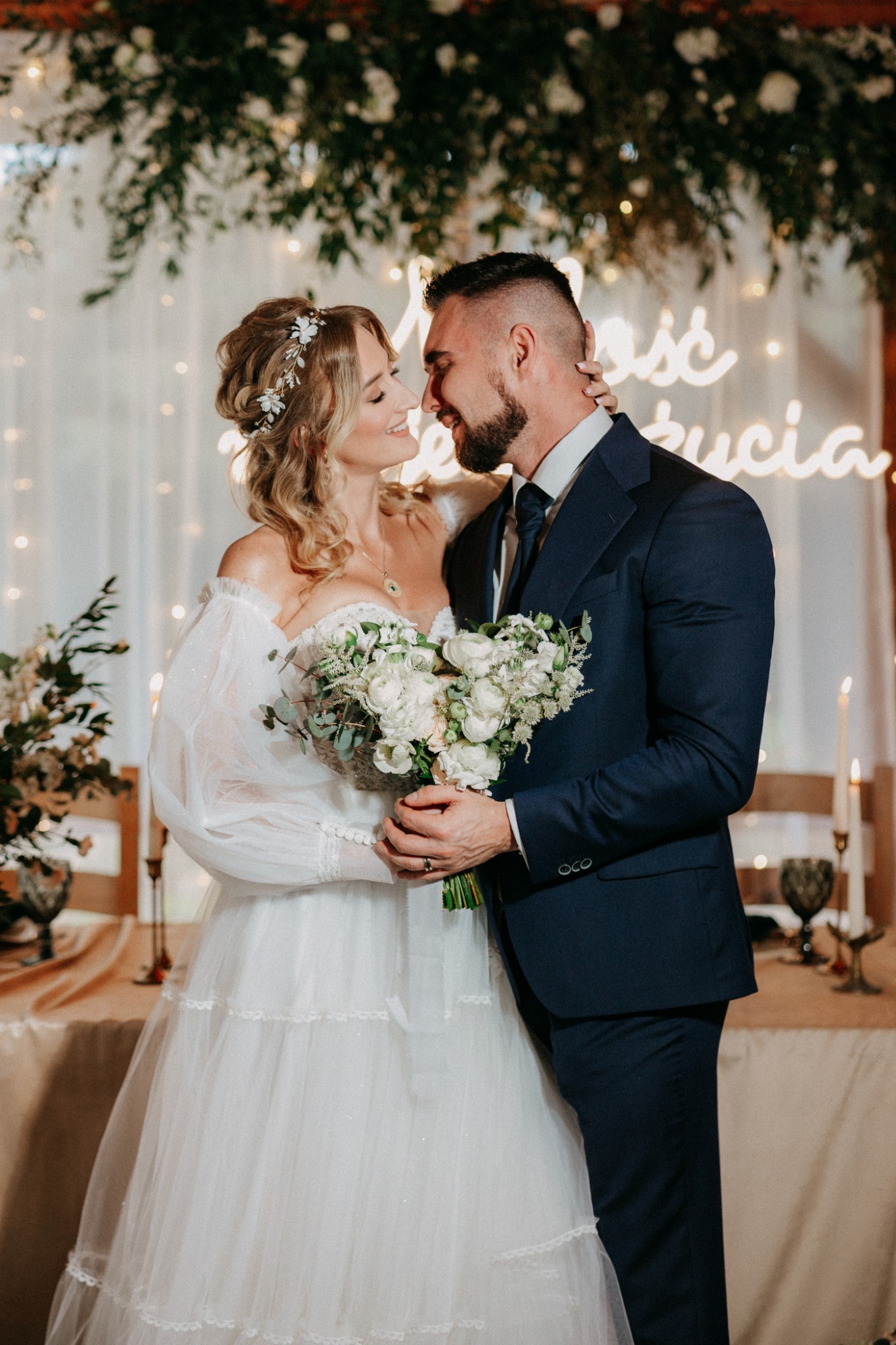 Para młoda w objęciach, panna młoda w białej sukni z bukietem, pan młody w granatowym garniturze, tło z girlandą świetlną i dekoracją kwiatową.