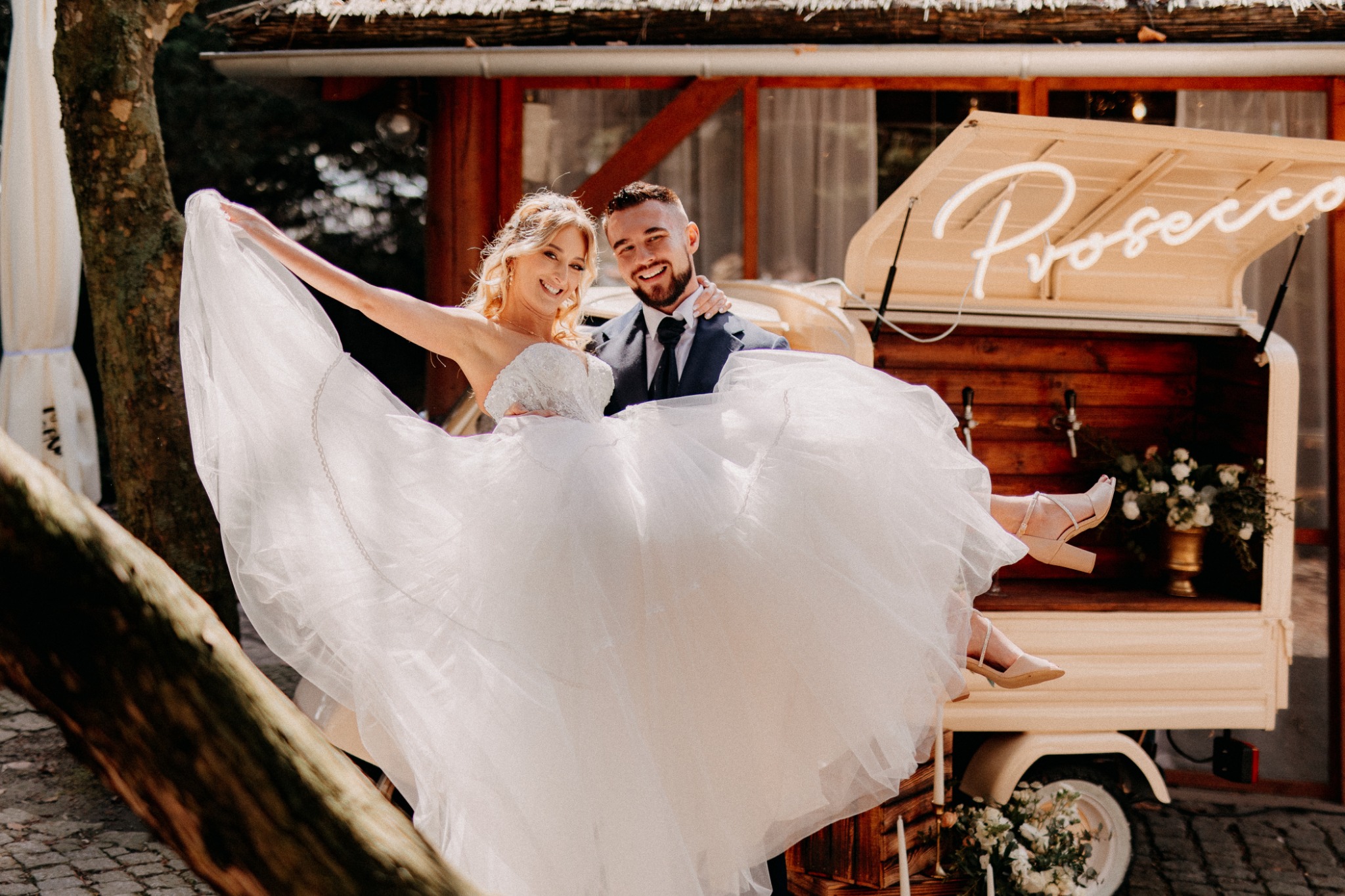 Uśmiechnięty pan młody trzyma na rękach pannę młodą w białej sukni na tle mobilnego baru z napisem 'Prosecco' i kwiatowymi dekoracjami.