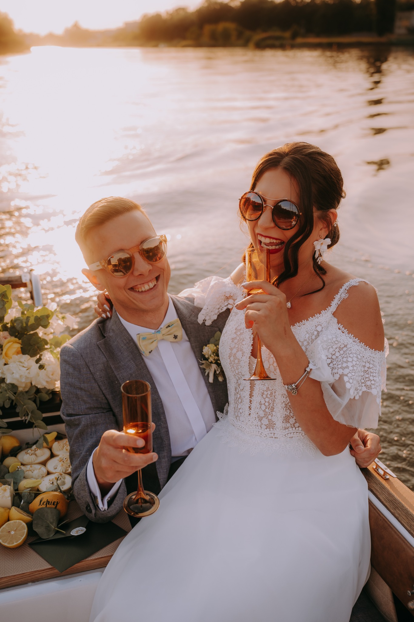 Uśmiechnięta para młoda w okularach przeciwsłonecznych, siedząca w łodzi z dekoracjami kwiatowymi i cytrynami, wznosząca toast złotymi kieliszkami na tle wody w promieniach zachodzącego słońca.
