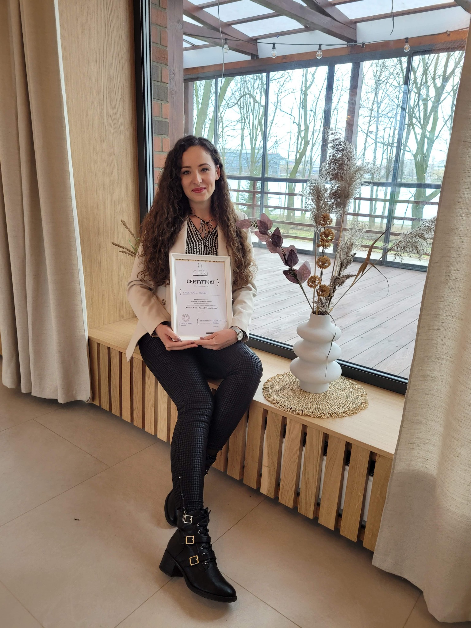Kobieta z kręconymi włosami siedzi na parapecie przy oknie, trzymając w rękach certyfikat ukończenia kursu Wedding Planner, obok wazon z suszonymi kwiatami.