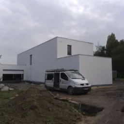 Nowoczesny, biały dom jednorodzinny z płaskim dachem, po świeżo wykonanej elewacji, z otwartymi drzwiami garażowymi i zaparkowanym vanem z otwartymi drzwiami bocznymi na błotnistym podjeździe.