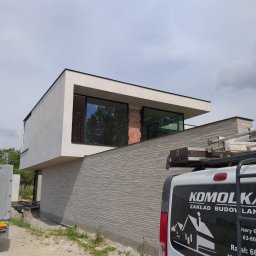 ZAKŁAD OGÓLNOBUDOWLANY Mariusz Komolka - Nowoczesny dom w trakcie wykańczania elewacji, widoczna część ściany z szarej cegły dekoracyjnej, okna z czarnymi ramami i biały fragment elewacji. Na pierwszym planie samochód dostawczy z logo...