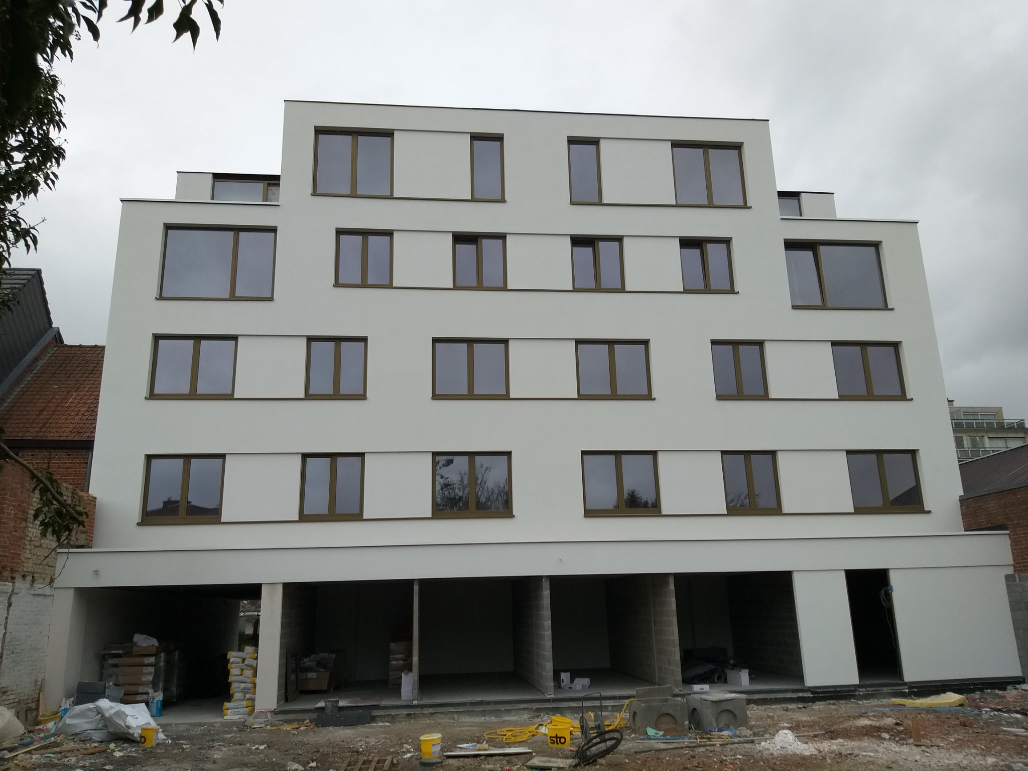 Nowoczesny budynek wielorodzinny z białą elewacją i brązowymi ramami okiennymi, widok z dołu na placu budowy z widocznymi materiałami budowlanymi i narzędziami.