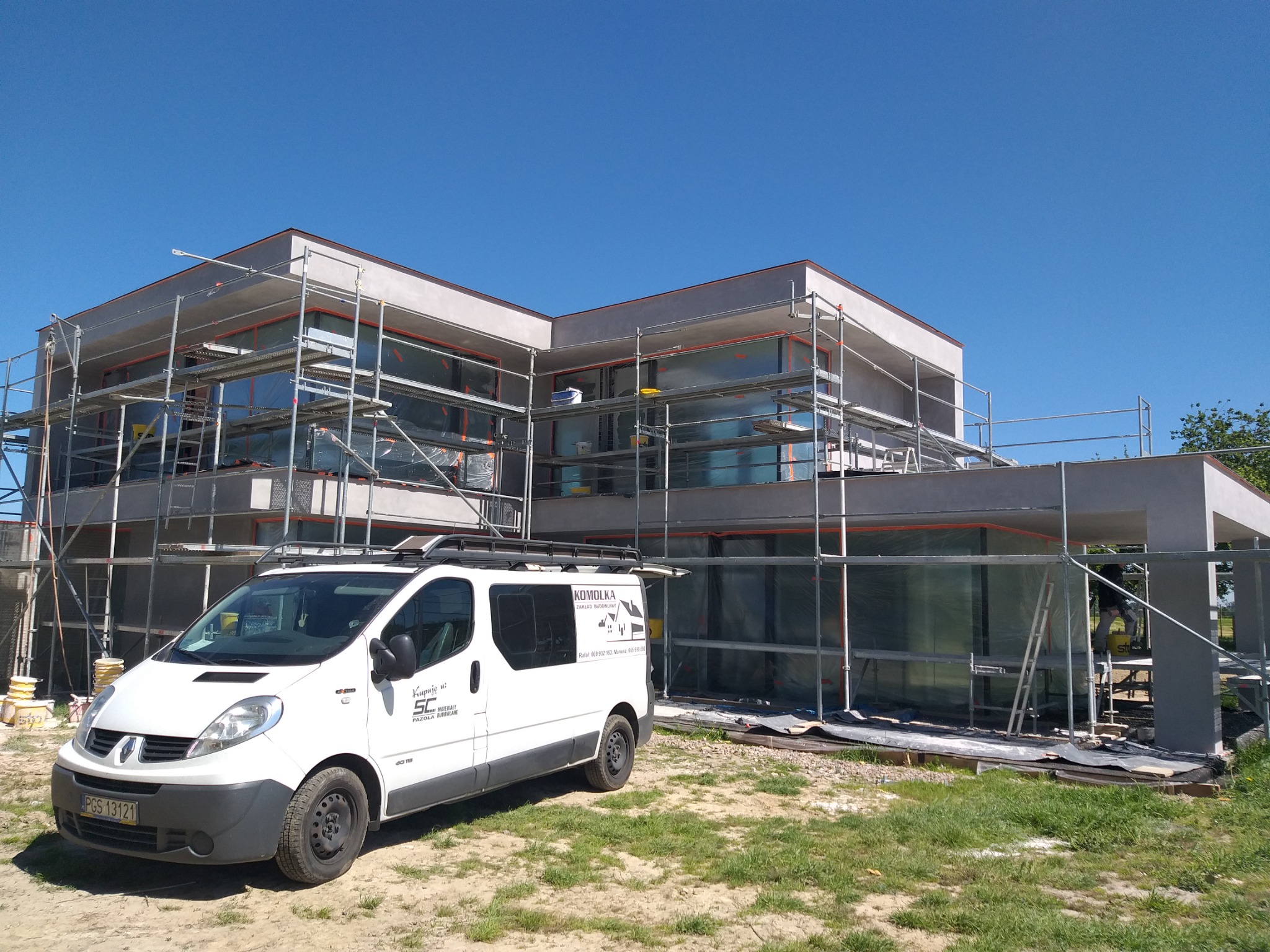 Nowoczesny dom w trakcie prac elewacyjnych, otoczony rusztowaniami, z zaparkowanym białym busem z logo firmy budowlanej na tle błękitnego nieba.