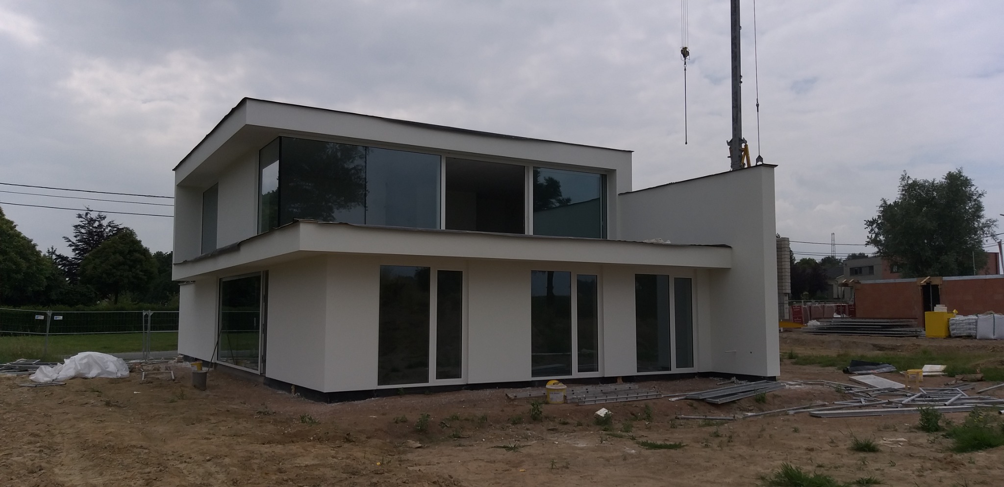 Nowoczesny, dwukondygnacyjny dom z płaskim dachem i dużymi oknami, świeżo po wykonaniu elewacji, z widocznym żurawiem budowlanym w tle.