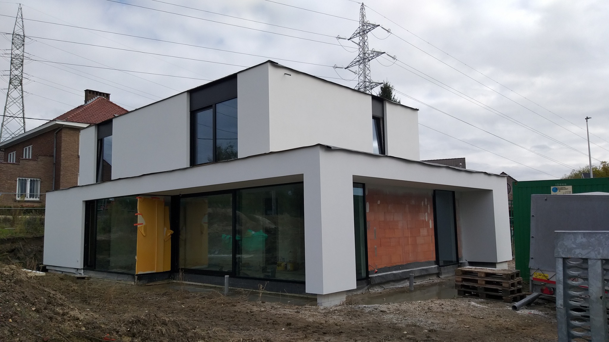 Nowoczesny, dwukondygnacyjny dom z płaskim dachem w trakcie budowy, z widoczną surową elewacją i dużymi oknami, na tle pochmurnego nieba i linii wysokiego napięcia.