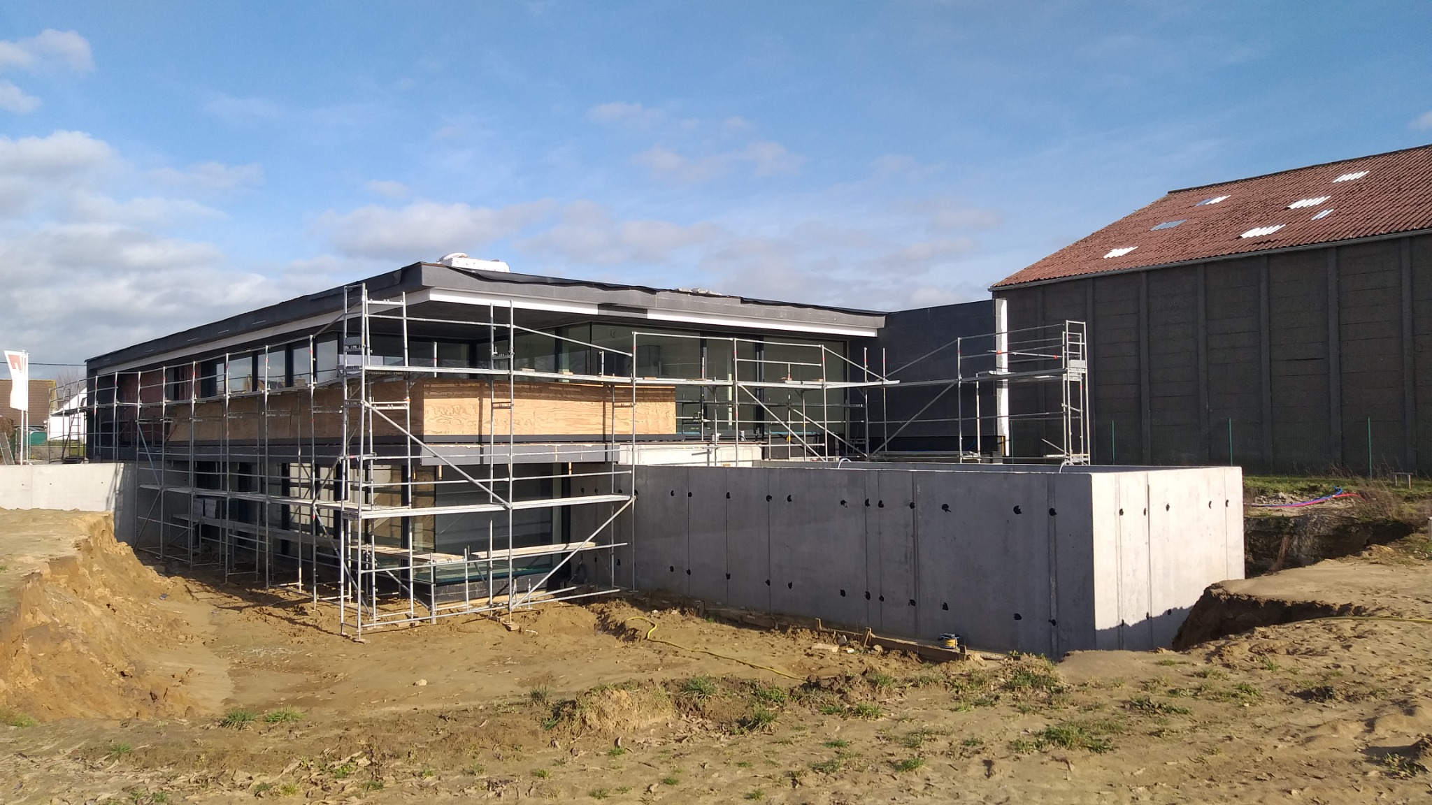 Budynek z płaskim dachem w trakcie ocieplania, widoczne rusztowania, płyty OSB i betonowa podmurówka z otworami wentylacyjnymi, obok budynek gospodarczy z czerwonym dachem.