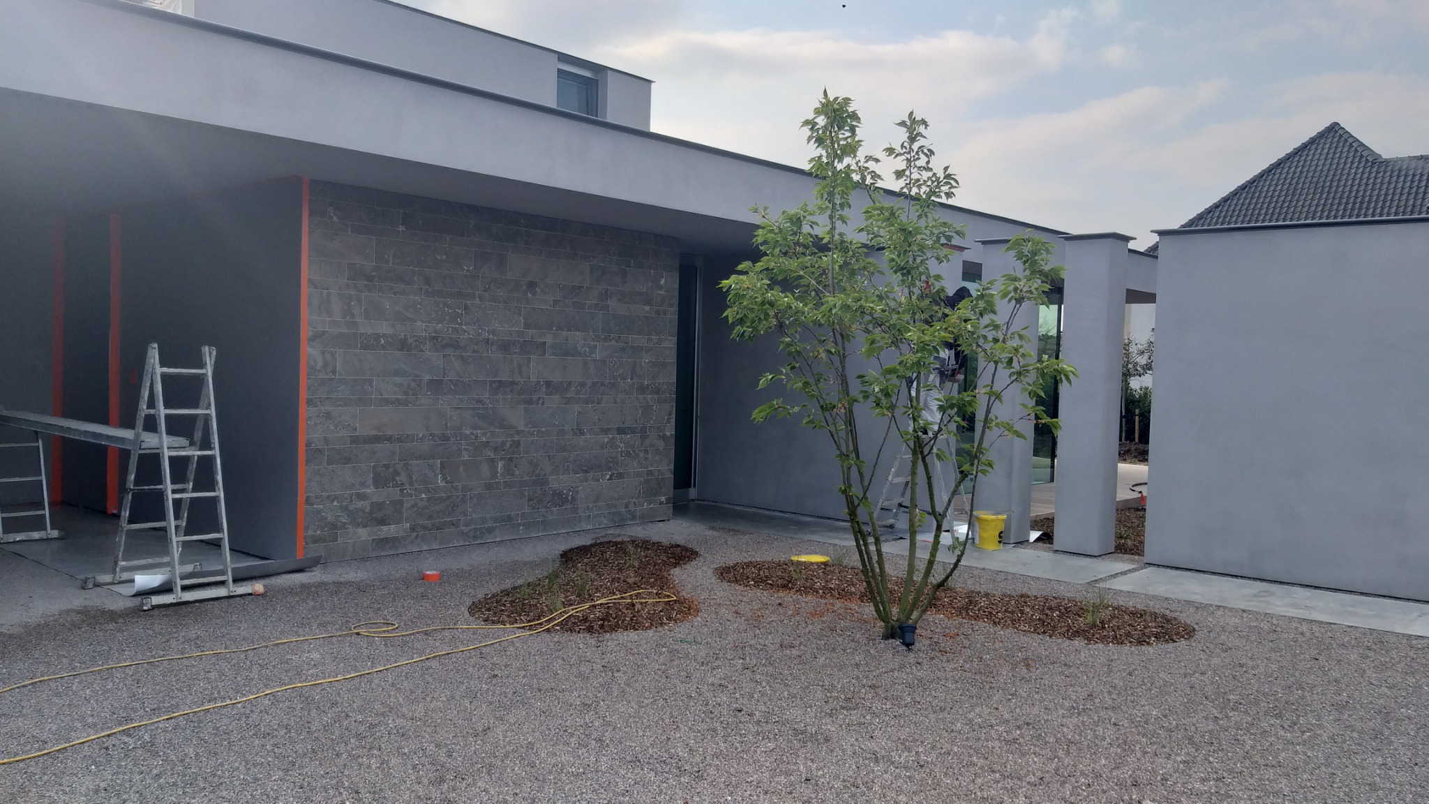 Nowoczesny dom z szarą elewacją z elementami imitującymi kamień, drabina malarska ustawiona przy ścianie, żółty kabel na żwirowym podłożu.