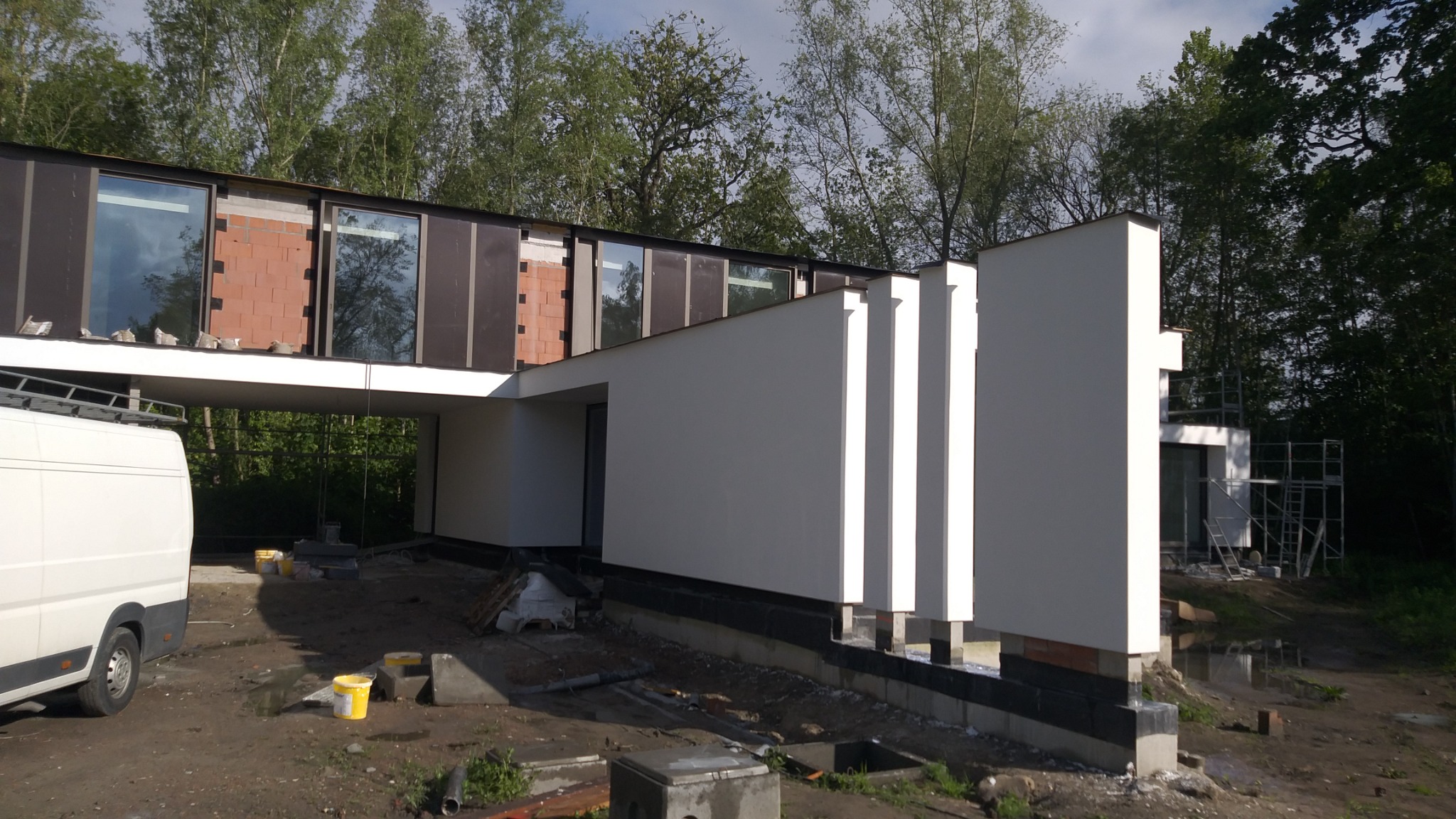 Nowoczesny dom w trakcie wykańczania elewacji, widoczne fragmenty cegieł i okna, na pierwszym planie częściowo widoczny biały van, teren budowy z elementami konstrukcyjnymi i żółtymi wiadrami.