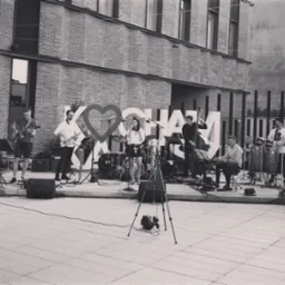 Zesp&oacute;ł muzyczny grający na zewnątrz, z saksofonistą, gitarzystą, wokalistką, kontrabasistą, klawiszowcem i perkusistą, na tle ceglanego budynku i dekoracji z napisem 'KOCHAM'